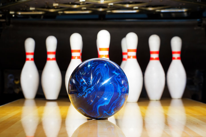 A blue bowling ball rolling toward a set of white pins with red stripes, reflecting on the polished wooden lane in a bowling alley.